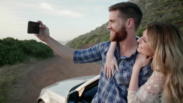 Couple-on-road-trip-taking-selfies-by-convertible