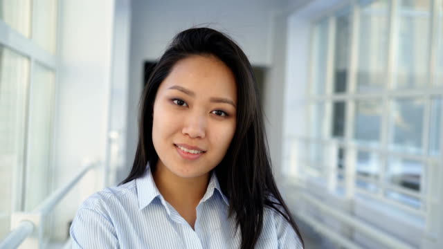 Portrait-of-young-beautiful-pretty-female-student-of-asian-ethnicity-standing-in-wide-white-hallway-indoors-looking-at-camera-and-smiling-positively