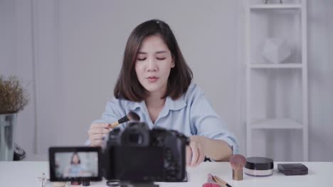 Feliz-sonriente-blogger-mujer-o-belleza-asiática-con-cepillo-y-cámara-de-grabación-de-video-y-agitando-la-mano-en-casa.-Concepto-de-belleza-videoblog-blog-personas.