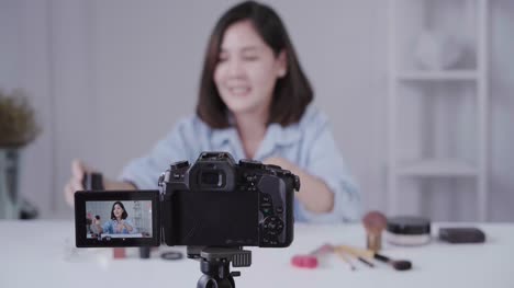 Glücklich-lächelnd-asiatische-Frau-oder-Beauty-Blogger-mit-Pinsel-und-Kamera-Videoaufnahme-und-winkenden-Hand-zu-Hause.-Videoblog-Blogging-Menschen-Schönheitskonzept.