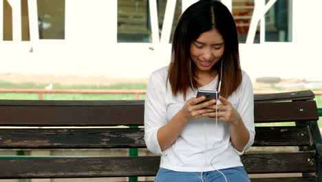Young-asian-woman-using-smartphone-sitting-on-a-bench-in-a-public-park.