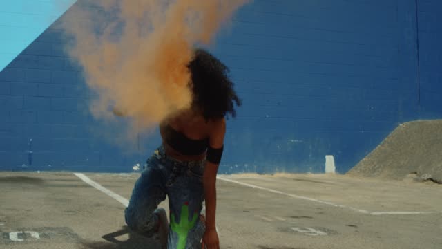 Beautiful-young-woman-holding-colorful-smoke-grenade-dancing-outside-against-blue-wall