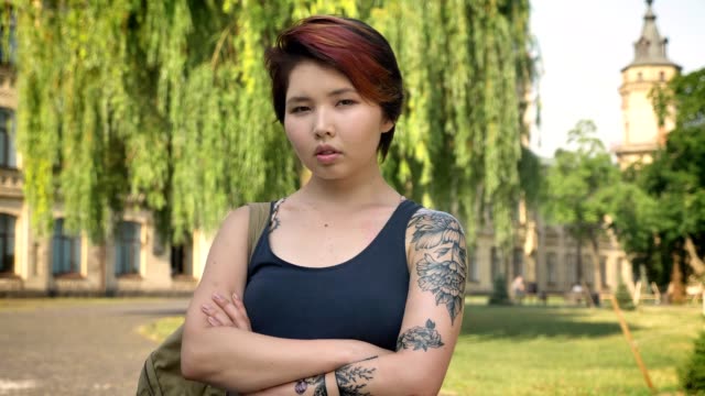 Portrait-of-young-asian-women-with-tattoo-standing-with-crossed-hands-and-looking-in-camera-in-park-near-university,-confident-and-serious