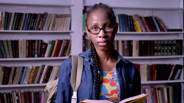 Young-pretty-african-american-woman-in-glasses-reading-book-in-library,-looking-at-camera,-serious-and-concentrated,-bookshelves-background