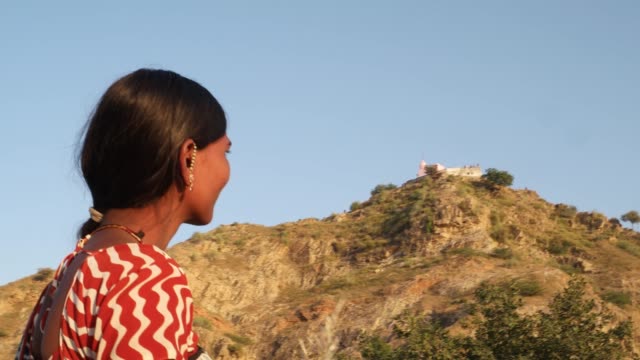 Pretty-young-Hindu-woman-in-ethnic-costume-waiting-and-looking-on-hill-top-Hindu-temple-and-contemplates-thinks-prays-for-a-visit-pilgrimage-saint-sacred-holy-religious-church-building-nature-outdoors