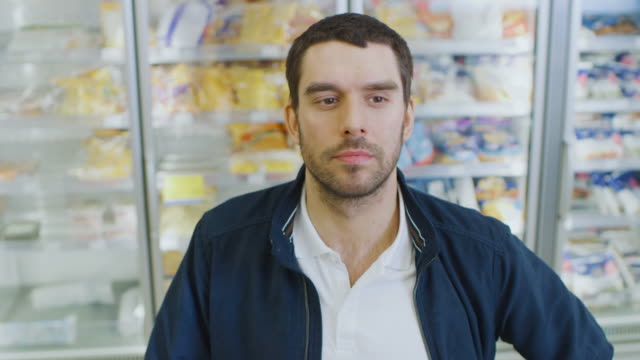 Portrait-Shot-of-the-Handsome-Man-Choosing--Tin-Can-from-the-Canned-Goods-Section-and-Places-it-In-His-Shopping-Cart.-In-the-Background-Frozen-Goods-Section-of-the-Store.