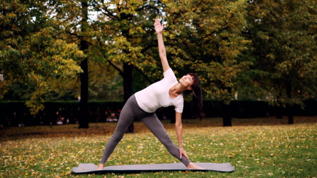 Attractive-young-woman-is-doing-yoga-outdoors-standing-on-mat-and-practising-sequence-of-asanas-Triangle-pose-and-Warrior-position-enjoying-fresh-air.