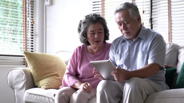 Senior-pareja-sujetando-el-tablet-y-sentados-en-el-sofá-juntos-en-la-sala-de-estar-en-casa.-Concepto-de-familia-feliz,-post-jubilación,-calidad-vida,-salud-mental-y-psicología-positiva.-resolución-de-4-k.