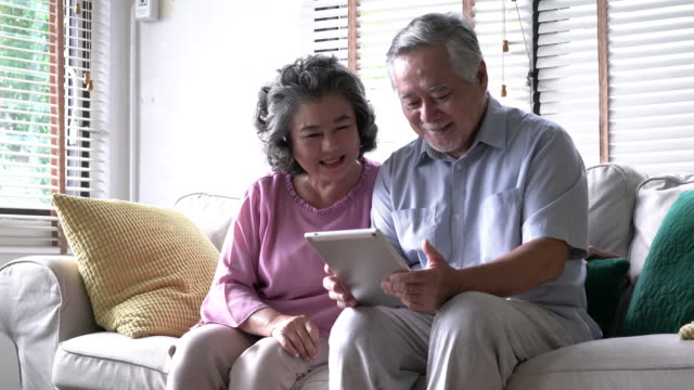 Paar-Senior-Tablet-halten-und-zusammensitzen-auf-Sofa-im-Wohnzimmer-zu-Hause.-Konzept-der-glücklichen-Familie,-Post-Ruhestand,-Lebensqualität,-psychische-Gesundheit-und-positive-Psychologie.-4-k-Auflösung.