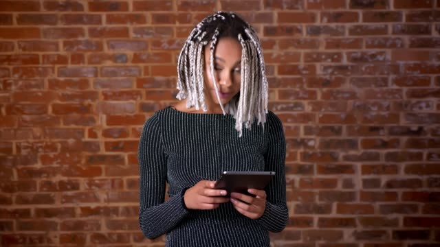 Young-African-girl-with-dreadlocks-using-a-tablet-computer-looking-at-the-camera-and-smiling,-Brick-wall-in-the-background.