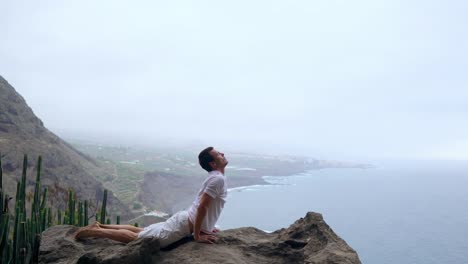 La-mujer-sentada-en-el-borde-de-un-acantilado-en-la-pose-del-perro-con-vistas-al-mar,-respirar-el-aire-de-mar-durante-un-viaje-del-yoga-a-través-de-las-islas