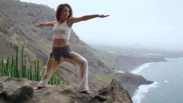 Young-woman-doing-yoga-in-the-mountains-on-an-island-overlooking-the-ocean-standing-on-one-leg-raising-her-hands-up