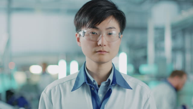 On-High-Tech-Factory:-Portrait-of-Asian-Worker-Wearing-Uniform-and-Safety-Goggles.-In-the-Background-Blurred-Electronics-Assembly-Line-with-Bright-Lights-and-other-Workers-Doing-their-Jobs.