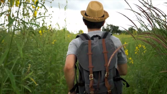 Back-view-of-young-Asian-male-tourist-backpacker-with-hat-and-bag-running-in-the-countryside-grassland-in-Thailand
