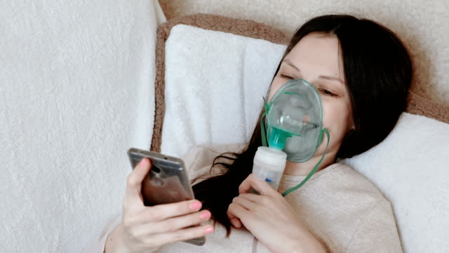 Verwenden-Sie-Vernebler-und-Inhalator-für-die-Behandlung.-Junge-Frau-Einatmen-durch-Inhalator-Maske-auf-der-Couch-liegen-und-im-Handy-Chat.-Seitenansicht.