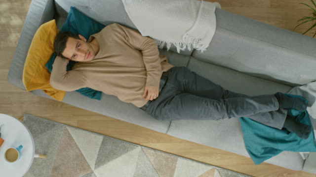 Sad-Young-Hispanic-Man-in-Brown-Jumper-and-Grey-Jeans-is-Lying-Down-on-a-Sofa.-Cozy-Living-Room-with-Modern-Interior-with-Plants,-Table-and-Wooden-Floor.-Top-View.