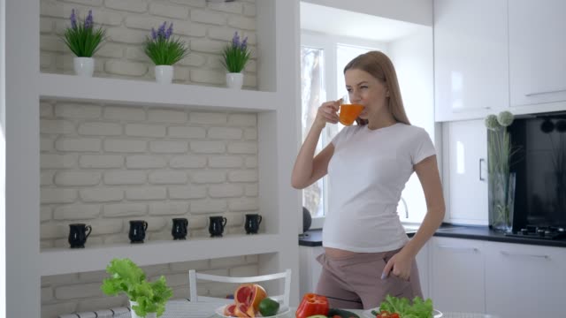 Retrato-de-feliz-maternidad-mujer-con-vientre-grande-beber-jugo-de-fruta-fresca-durante-el-almuerzo-saludable-en-la-cocina-en-Apartamento