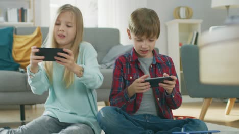At-Home-Sitting-on-a-Carpet:-Cute-Little-Girl-and-Sweet-Boy-Playing-in-Competitive-Video-Game-on-two-Smartphones,-Holding-them-in-Horizontal-Landscape-Mode.-Close-up-Portrait-Camera-Shot.
