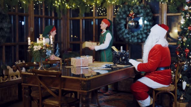 Santa-Claus-schreibt-einen-Brief-an-die-Schreibmaschine-und-beobachten-wie-die-Elfen-gegenseitig-anschubsen-präsentiert-vor-dem-Hintergrund-der-Weihnachtsschmuck-in-den-Raum-am-Vorabend-des-neuen-Jahres
