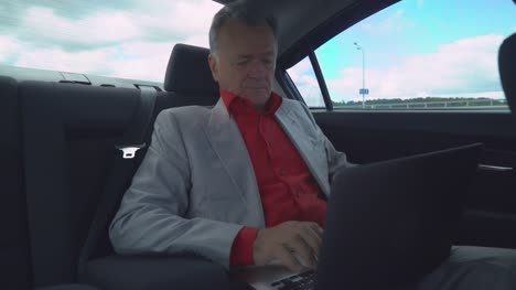 Hombre-envejecido-medio-utilizando-la-computadora-portátil-en-el-coche-se-sienta-en-el-asiento-trasero
