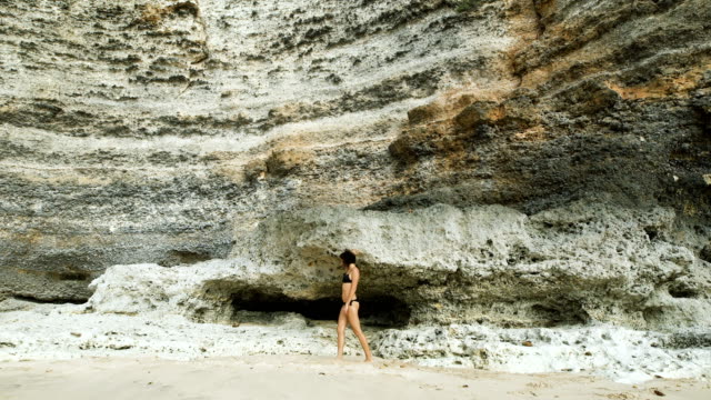 Una-chica-en-bañador-en-la-playa.-Contra-el-fondo-de-un-enorme-muro-de-piedra.-Ella-camina-a-lo-largo-de-la-playa.-Camina-hacia-adelante-y-hacia-atrás.-Bali