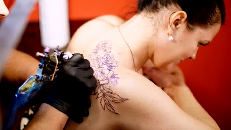close-up,-in-a-tattoo-salon-,-a-specialist-is-doing-a-tattoo-on-woman's-back,-black-paint-floral-ornament.-a-man-works-in-special-black-gloves,-on-special-equipment
