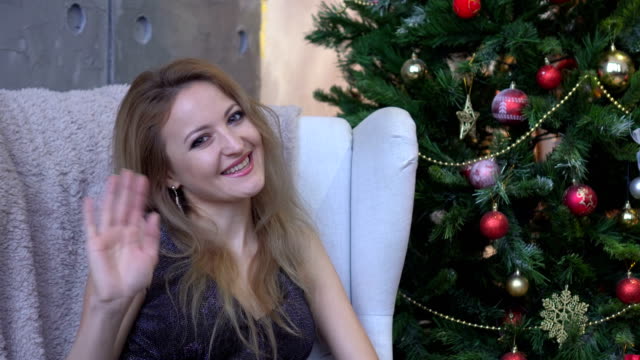 Eine-schöne-lächelnde-Frau-in-einem-glänzenden-Kleid-winken-und-sitzen-auf-einem-Stuhl-in-der-Nähe-von-einem-Weihnachtsbaum
