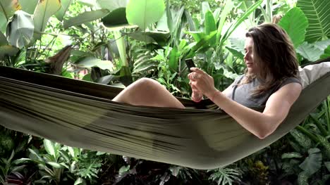 Woman-using-smartphone-while-sitting-on-hammock,-exotic-background