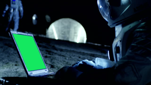 Astronauta-del-planeta-extranjero-trabaja-en-un-portátil-con-pantalla-verde-de-maqueta.-En-el-fondo-su-miembro-de-la-tripulación-y-los-Habitat-del-espacio.-Concepto-de-colonización-extraterrestre.