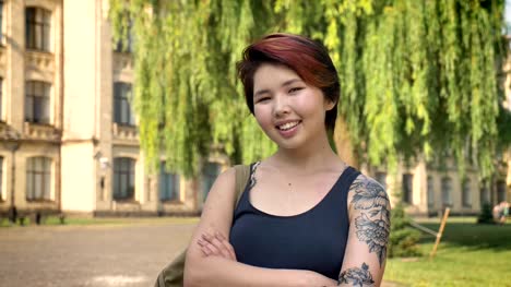 Portrait-of-young-asian-women-with-tattoo-standing-with-crossed-hands-and-looking-into-camera-in-park-near-university,-smiling,-happy