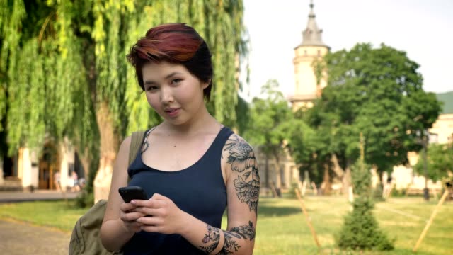 Asiatische-junge-tätowierte-Frauen-stehen-und-SMS-auf-Handy-im-Park-in-der-Nähe-von-Universität,-Lächeln,-glücklich