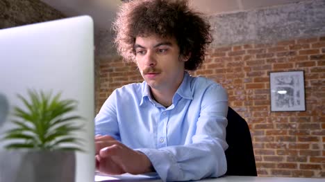 Lustige-Nerd-Programmierer-mit-dem-lockigen-Haar-Codierung-auf-Laptop-und-sitzen-am-Tisch-im-modernen-Büro
