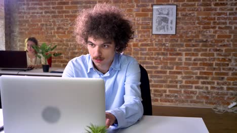 Funny-nerd-programmer-with-mustache-and-curly-hair-coding-on-laptop-sitting-at-table,-modern-office-background
