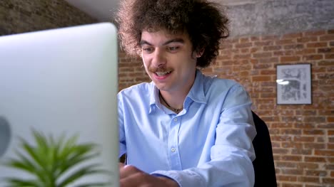 Lustige-junge-Büroangestellte-mit-lockigem-Haar-auf-Laptop-tippen-und-sitzen-am-Tisch-im-modernen-Büro,-Lächeln