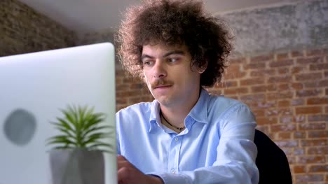 Lustige-nerdy-Mann-mit-lockiges-Volumen-Haar-denken-und-tippen-auf-Laptop,-sitzen-im-modernen-Büro,-konzentriert-und-entschlossen