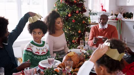 Mixed-race,-multi-generation-family-sitting-at-their-Christmas-dinner-table-reading-jokes-and-putting-on-paper-party-hats-from-Christmas-crackers,-close-up
