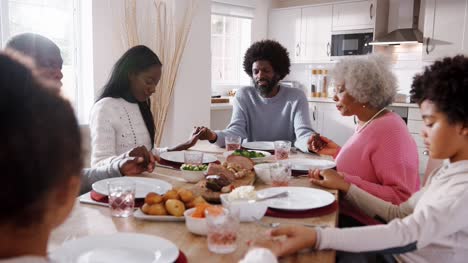 Multi-generation-mixed-race-family-holding-hands-and-saying-grace-at-the-table-before-eating-their-Sunday-dinner,-selective-focus