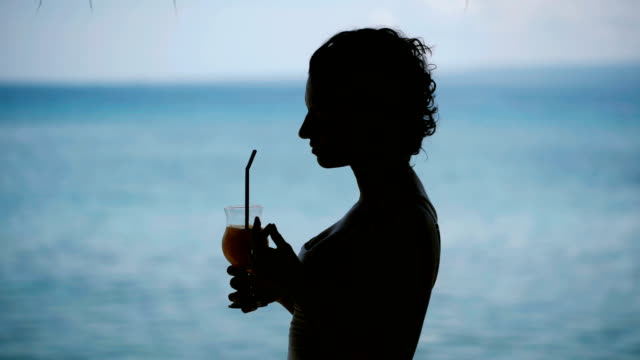 brunette-with-short-hair-standing-near-the-seaside-on-the-island-of-Bali.-She-is-very-focused,-looking-away-and-drinking-a-cocktail.-Wonderful-weather,-the-wind-and-the-island-promotes-tranquility-and-meditation-tourists