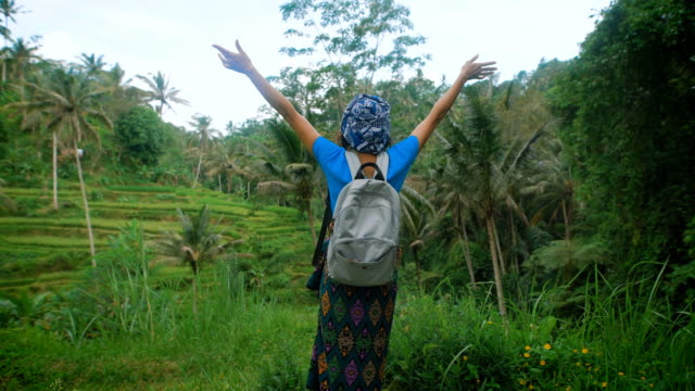 Viajero-femenino-ruso-vestido-con-ropa-azul-casual-es-de-pie-en-medio-de-campos-sin-fin-Indonesia,-levanta-las-manos-y-disfrutando-de-una-frondosa-vegetación-tropical-y-aire-fresco-alrededor-de