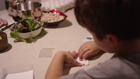 Ein-kleiner-Junge-setzt-einen-weihnachtliche-Sticker-auf-ein-Stück-Papier,-während-seine-Mutter-essen-bereitet
