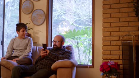 Un-padre-y-un-hijo-sentarse-y-hablan-sobre-un-sofá-en-casa-mientras-que-el-padre-utiliza-su-teléfono