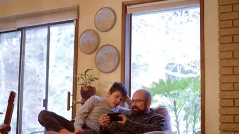 Vater-und-Sohn-sitzen-und-sprechen-auf-einer-Couch-zu-Hause,-während-der-Vater-sein-Handy-benutzt