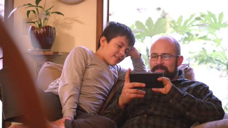 A-father-and-son-sit-and-talk-on-a-couch-at-home-while-the-dad-uses-his-phone