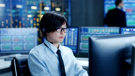 In-the-Network-Operations-Center-Trader-Makes-Personal-Client-Call-with-a-Headset.-He's-Surrounded-by-Monitors-Showing-Stocks-Data-and-Graphs.