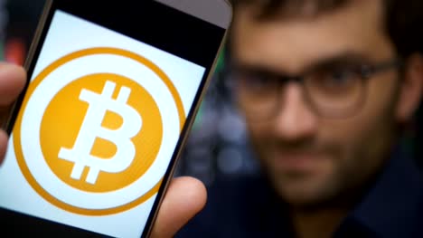 Man-earns-bitcoins-on-his-bitcoin-farm.-Male-making-a-payment-with-bitcoins-cryptocurrency-using-his-smartphone.-Custom-application-interface-design.-Guy-smiles,-earns-on-the-financial-market