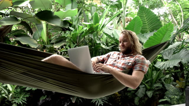 Hombre-trabajando-en-ordenador-portátil-mientras-está-sentado-en-la-hamaca,-fondo-exótico