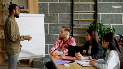 Young-african-american-businessman-explaining-start-up-business-plan-on-flipchart-with-blockchain-technology-presentation-to-colleagues-in-modern-loft-office-indoors