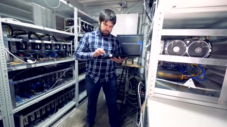 Male-programmer-is-standing-in-the-middle-of-a-mining-farm-unit-with-a-keyboard-and-working
