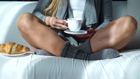 Mujer-sentada-con-la-taza-de-café-y-croissant-en-sofá-4K
