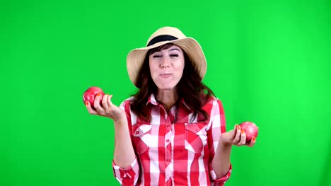 funny,-smiling-female-farmer-in-checkered-shirt-and-hat,-holding-in-hands-two-large-sour-apples,-eats,-bites-them,-wrinkles-her-face,-on-green-background-in-studio.-Healthy-food-to-your-table